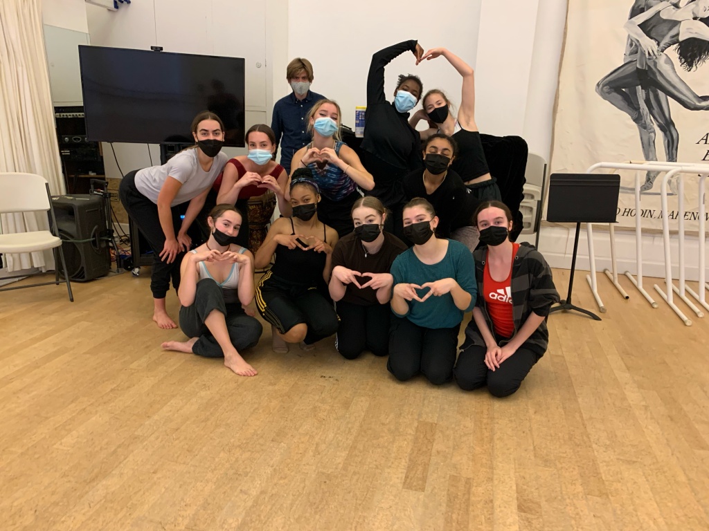 BFA at Dominican students gathered together with Amanda Harris in a studio at LINES Dance Center. All the dancers and the musician are wearing masks, and some of the students are creating hearts with their hands and arms.