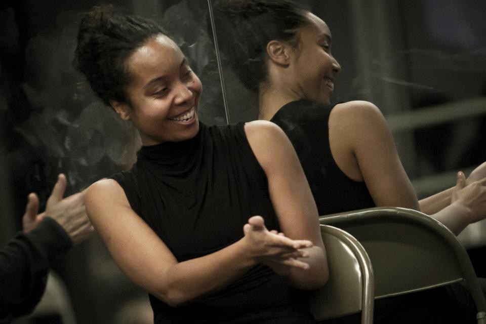 Sidra Bell sitting in a studio, smiling with her back to a mirror that shows her reflection