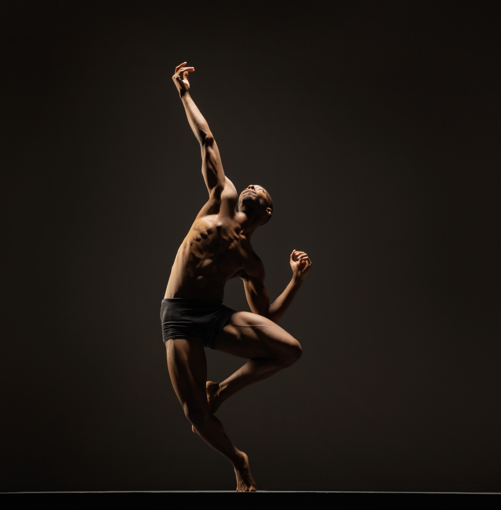 LINES Ballet company artist Joshua Francique with leg in passé, arm reaching up above head, focus directed toward the extended hand
