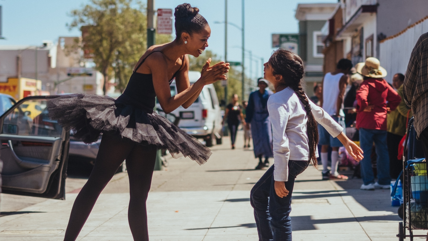Aesha Ash on a street in Rochester, New York, smiling and interacting with a smiling child who is motion; Aesha is wearing a black tutu, black tights, and a black leotard