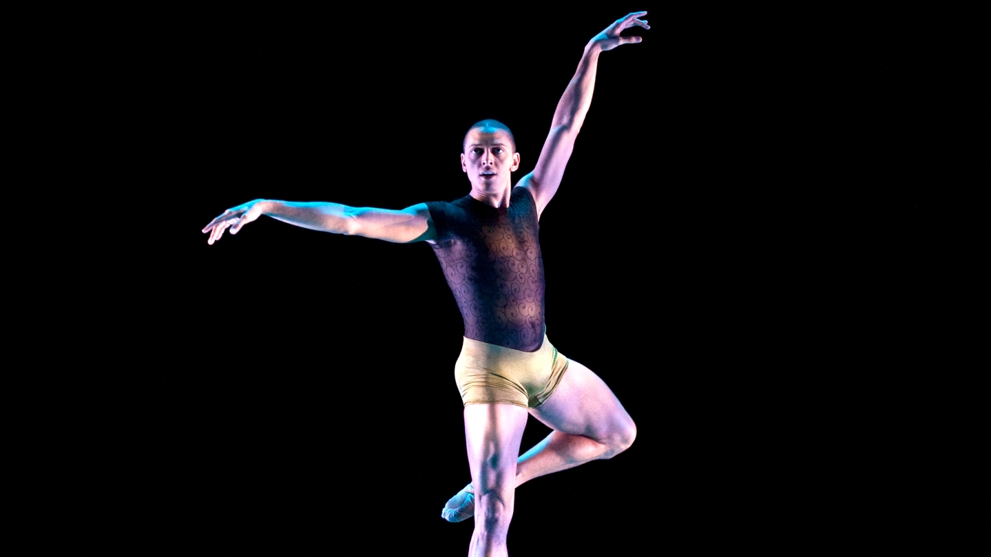 Clifton Brown performing Alonzo King's "Following the Subtle Current Upstream" with Alvin Ailey American Dance Theater; Clifton is on relevé with his other leg in passé; he is looking out with one arm extended above his head and the other extended out to the side