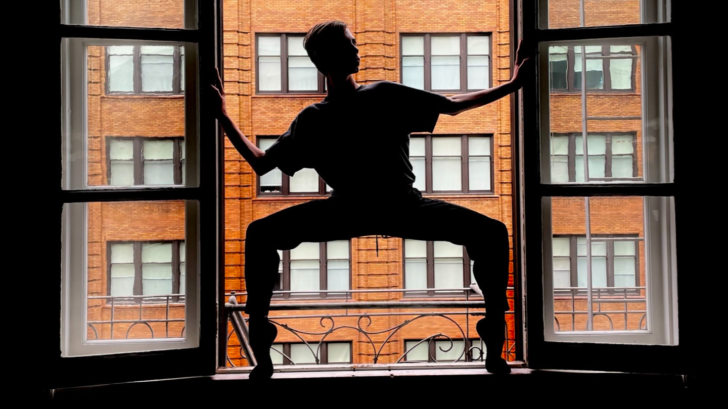 LINES Ballet Summer Program student Gabriel Warren dancing in a window; Gabriel is in a second position in relevé, holding the edges of the window